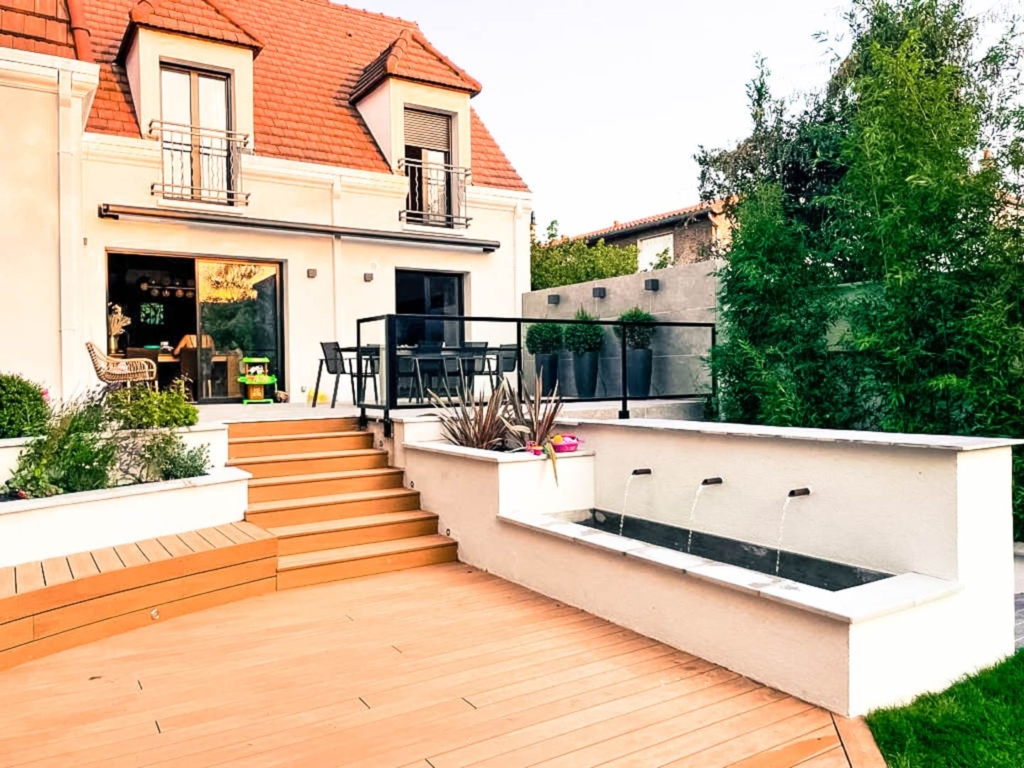 maconnerie-paysagere-bassin Terrasse en bois donnant sur des marches menant à un bassin ornemental avec trois fontaines murales, entourée de plantations et d’un espace repas extérieur.