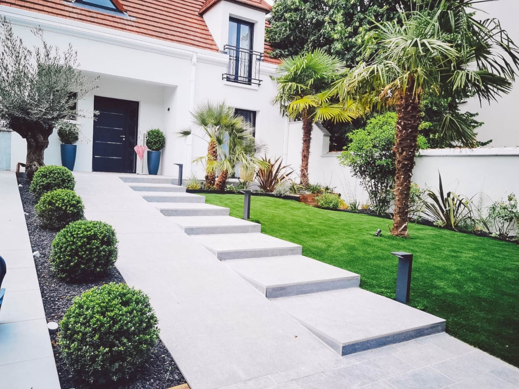 maconnerie-paysagere-escalier Entrée extérieure moderne avec escalier en béton, pelouse impeccable, massifs de buissons taillés et palmiers créant une ambiance végétale soignée.