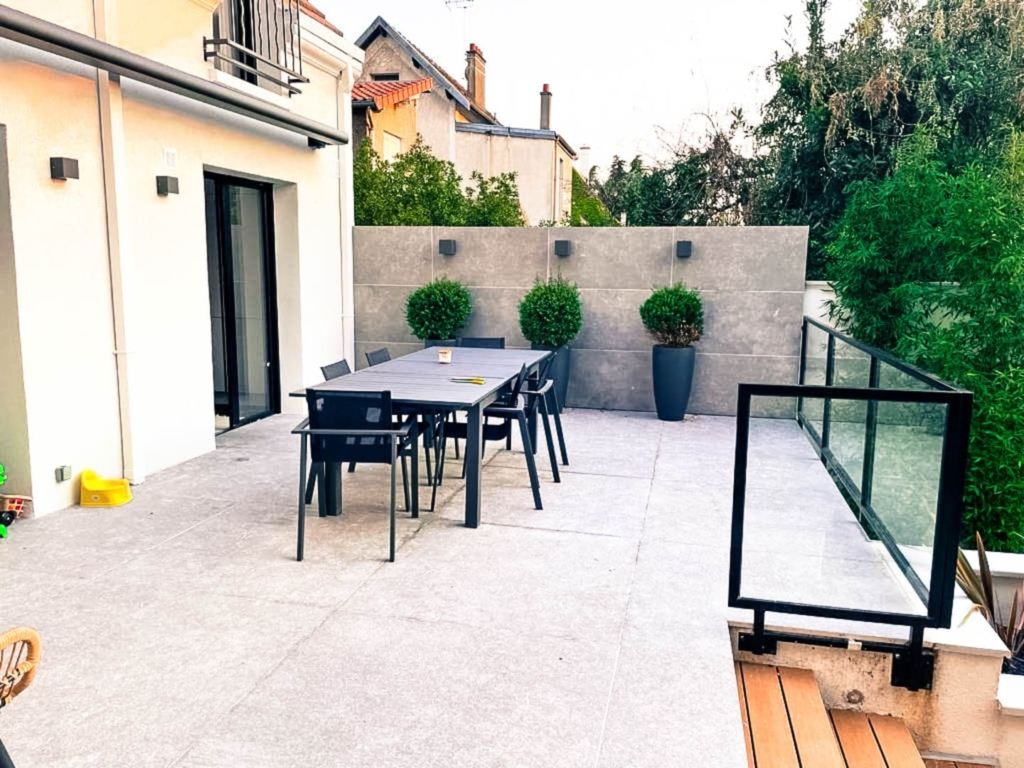 maconnerie-paysagere-terrasse-carrelee Grande terrasse carrelée attenante à la maison, équipée d’une table de repas extérieure et bordée de panneaux décoratifs et plantes en pots.