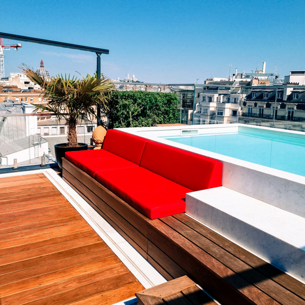 lmdj-amenagement-toit-terrasse-paris-4 Toit-terrasse à Paris avec piscine design, banquette en bois à coussins rouges et vue sur les toits parisiens sous un ciel bleu.