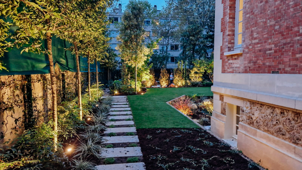 lmdj-jardin-luxe--2-paris Allée en dalles traversant un jardin paysager moderne, bordé d’arbres et de végétation éclairée au pied d’une maison en briques à Paris.