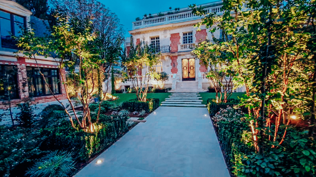 lmdj-jardin-luxe-3-paris Allée pavée illuminée menant à une élégante maison en pierre et briques, entourée d’un jardin paysager verdoyant en soirée.