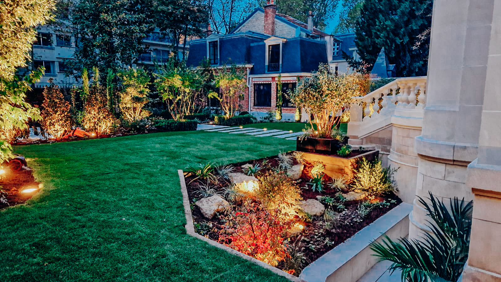 Jardin paysager haut de gamme avec pelouse soignée, massif fleuri éclairé et escalier en pierre menant à une maison en briques à Paris.