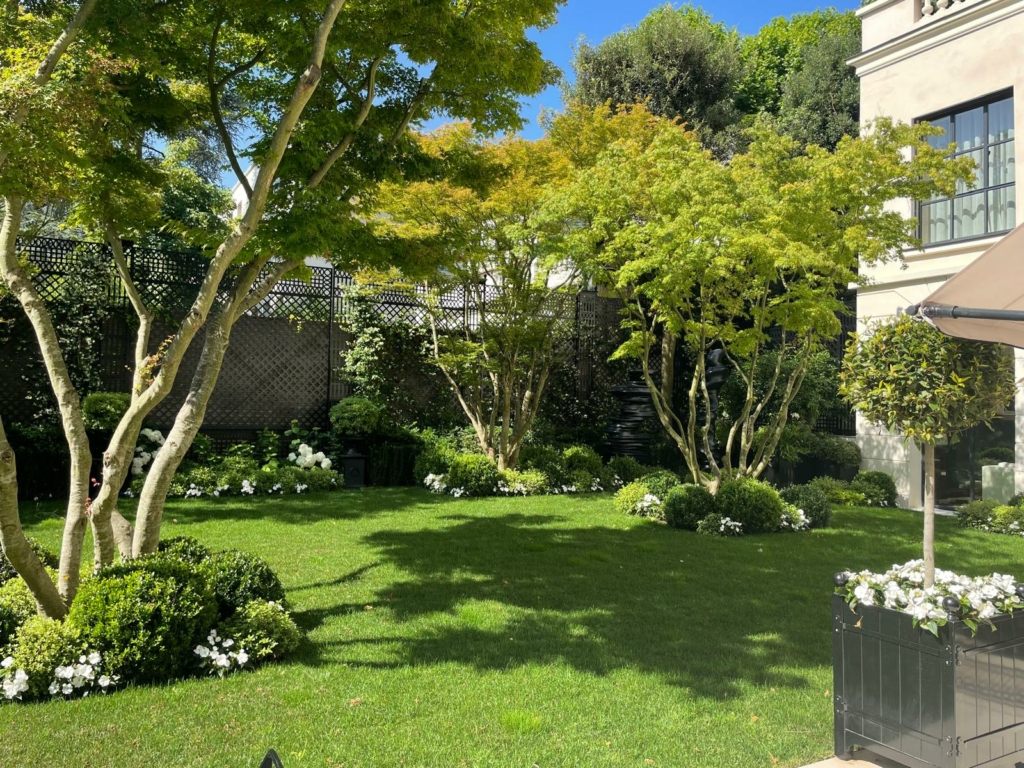 Jardin paysager à Paris avec pelouse verdoyante, massifs fleuris et façade d’hôtel particulier.