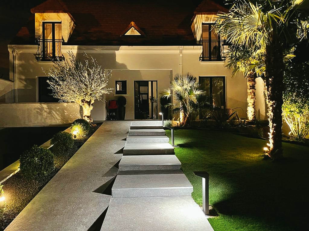 Allée design éclairée menant à une maison de prestige entourée de palmiers et d’oliviers, vue de nuit.