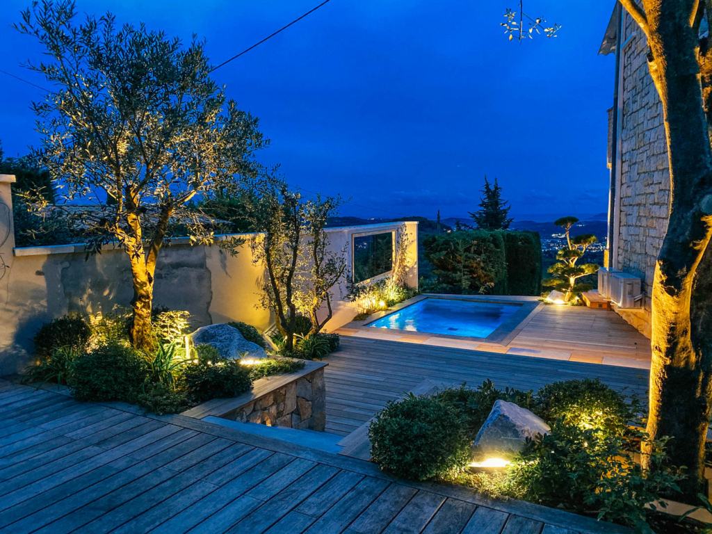Piscine design éclairée de nuit sur terrasse en bois, entourée d’oliviers et de végétation.