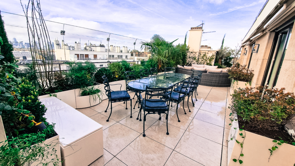 Terrasse en pierre naturelle avec table en verre, végétation luxuriante et vue sur les toits.