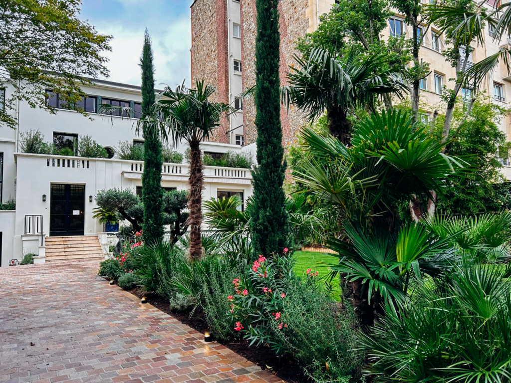 Allée pavée bordée de palmiers, cyprès et massifs fleuris devant une maison contemporaine.