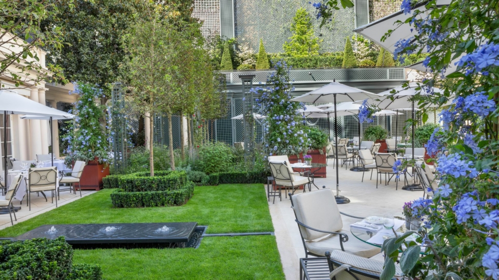 Terrasse gastronomique d’hôtel paysagée avec espaces de restauration, végétation soignée, pelouse structurée et fontaine décorative.