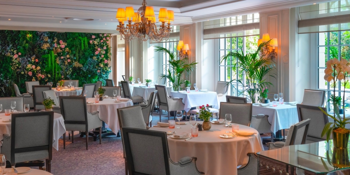 Salle du restaurant La Bauhinia au Shangri-La Paris en décor d'été, avec un mur végétal fleuri, des palmiers en bacs devant les baies vitrées et des tables dressées