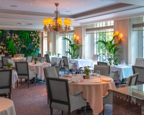 shangri-la-paris-2048x1366 (1) Salle du restaurant La Bauhinia au Shangri-La Paris en décor d'été, avec un mur végétal fleuri, des palmiers en bacs devant les baies vitrées et des tables dressées